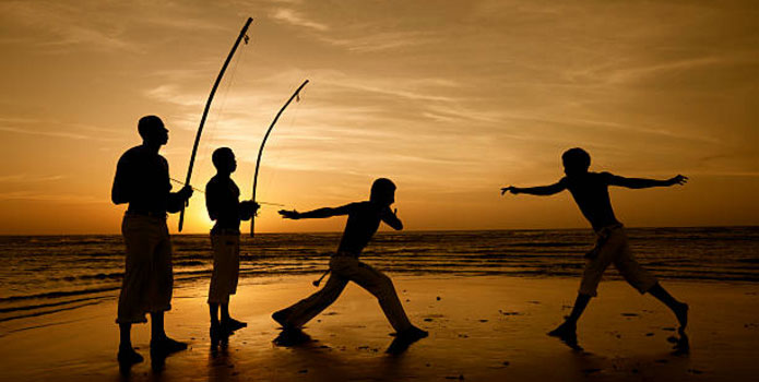 Four people play Capoeira, a Brazilian sport/martial-art/dance on the beach at sunset.